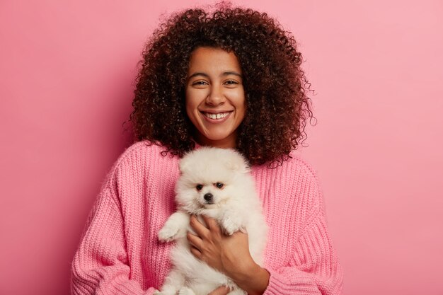 Mulher afro-americana positiva posa com spitz fofo nas mãos, cão de estimação, tem expressão alegre para adotar animais domésticos isolados sobre fundo rosa.