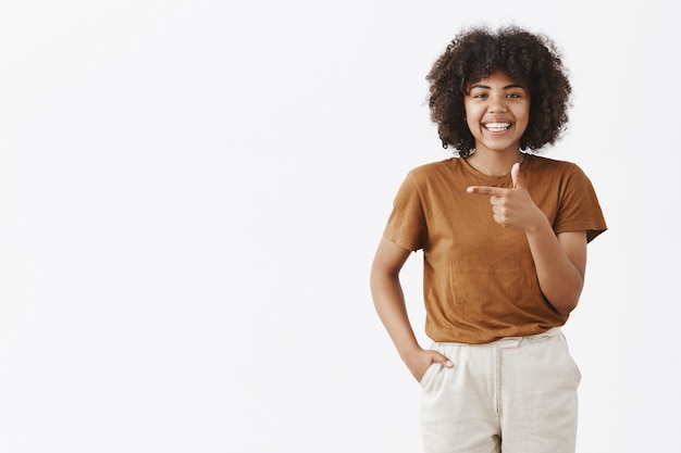 Mulher afro-americana satisfeita e feliz em uma camiseta marrom estilosa e calça segurando a mão no bolso, sorrindo alegremente enquanto aponta para a esquerda, dando conselhos para onde ir ou que caminho escolher ao invés da parede cinza