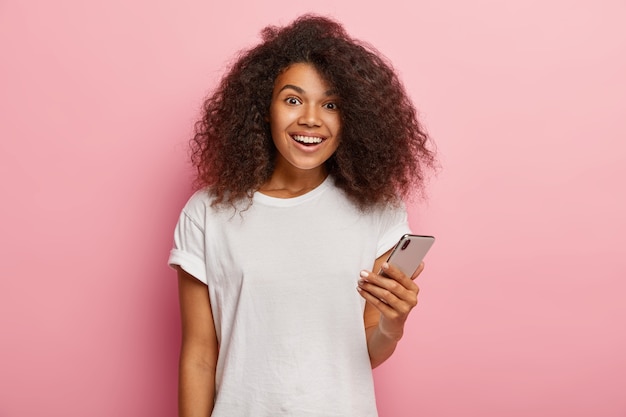 Mulher afro-americana satisfeita em trajes casuais, segura um smartphone moderno, espera a ligação e fica feliz em receber mensagem do namorado