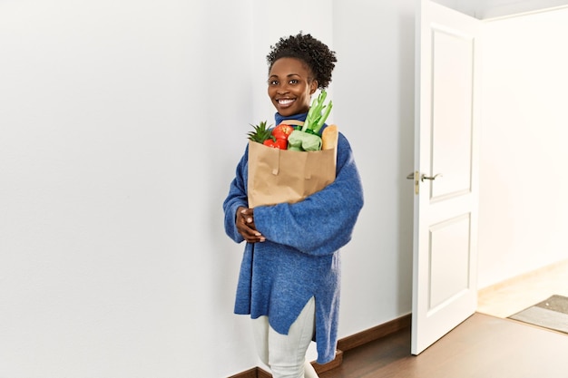 Mulher afro-americana segurando sacola de compras entrando pela porta em casa