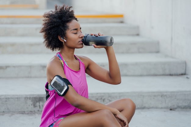 Mulher afro-atlética bebendo água e relaxando depois do treino enquanto está sentada na escada ao ar livre