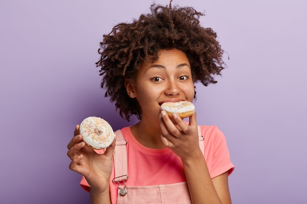 Mulher afro com fome morde rosquinha saborosa com granulado brilhante, tem nutrição prejudicial à saúde, não consigo imaginar a vida sem sobremesas doces, tem cabelo encaracolado, não mantém dieta, isolado na parede roxa