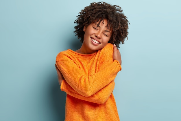 Mulher afro linda e charmosa fica de olhos fechados, sorri de prazer, mostra dentes brancos sente conforto, se abraça, usa blusão laranja, inclina a cabeça, modela sobre parede azul, tem autoestima elevada