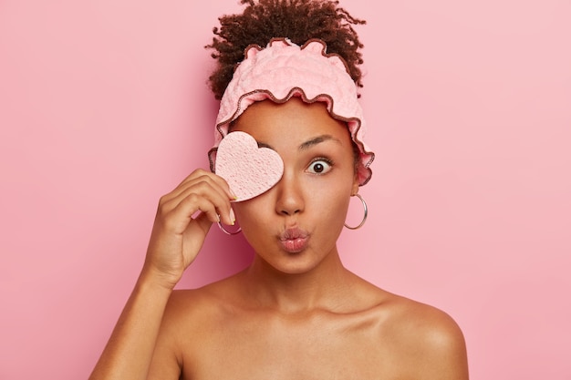 Foto grátis mulher afro surpresa cobre um olho com uma esponja cosmética, mantém os lábios arredondados, olhos esbugalhados, faz tratamentos de beleza no spa, penteava os cabelos cacheados