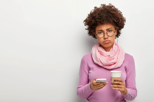 Mulher afro triste e descontente, cansada de trabalhar duro no escritório, segura o celular, baixa um novo aplicativo, segura o café para viagem, usa grandes óculos redondos, gola alta com lenço