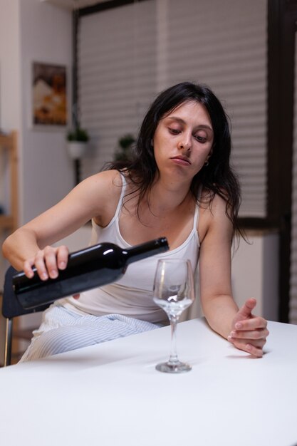 Mulher alcoólatra com uma garrafa de vinho e um copo, sentindo-se triste em casa. Pessoa solitária bebendo bebida com álcool, sendo depressiva. Adulto com vício sentindo-se emocional e chateado