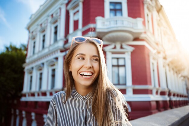 Mulher alegre bonita jovem em óculos de sol andando pela cidade, sorrindo.