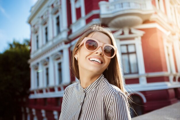 Mulher alegre bonita jovem em óculos de sol andando pela cidade, sorrindo.