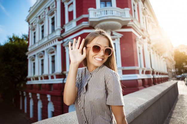 Mulher alegre bonita jovem em óculos de sol andando pela cidade, sorrindo.