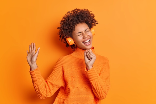 Foto grátis mulher alegre canta a música e mantém a mão perto da boca como se o microfone ouvisse sua playlist favorita por meio de fones de ouvido vestida de maneira casual em uma vívida parede laranja