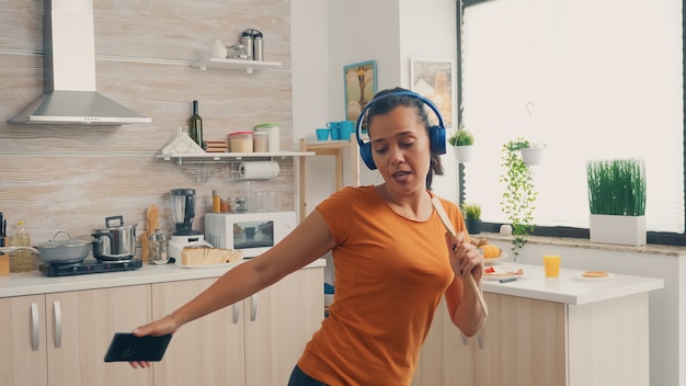 Mulher alegre cantando na colher de pau enquanto ouve música em fones de ouvido pela manhã. Dona de casa enérgica, positiva, feliz, engraçada e fofa dançando sozinha na casa. Entretenimento e lazer sozinho