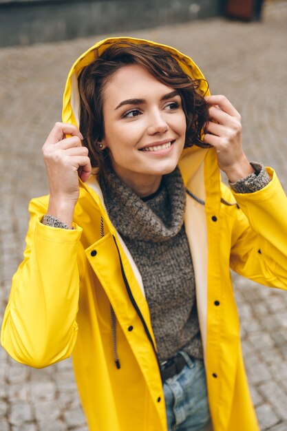 Mulher alegre caucasiana em capa de chuva amarela usando capuz e aproveitando o tempo enquanto caminhava no parque da cidade