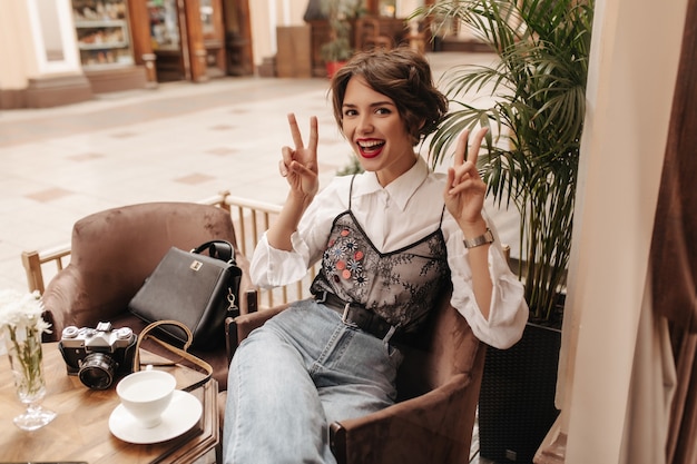 Mulher alegre com batom brilhante em jeans com cinto, mostrando sinais de paz no café. Mulher legal com cabelo curto em sorrisos de camisa branca no restaurante.