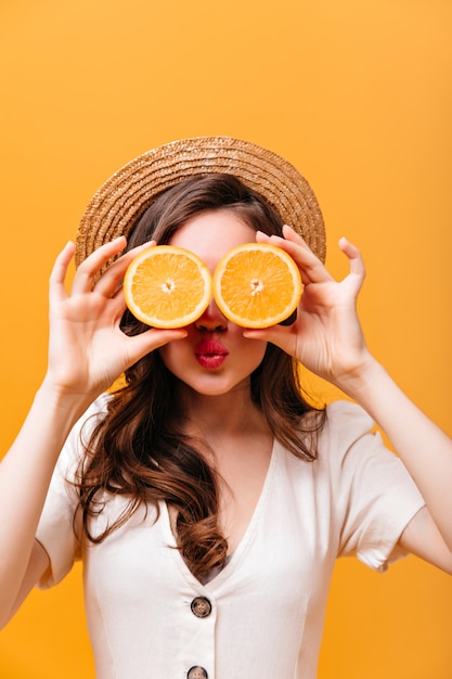 Mulher alegre com cabelos ondulados cobre os olhos com laranjas. senhora de velejador e camisa posando em fundo isolado.