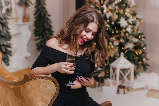 Mulher alegre com mensagem de texto de cabelo muito brilhante na noite de ano novo. Retrato interior de menina na moda de vestido preto bebe champanhe e segurando o telefone perto da árvore de Natal.