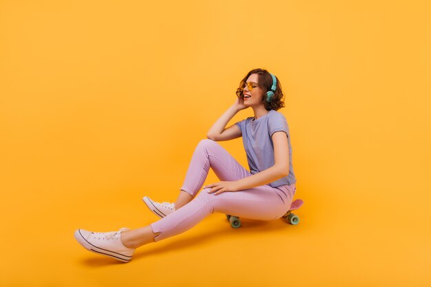 Mulher alegre com roupa elegante, sentada no skate com os olhos fechados. Garota maravilhosa de óculos amarelos, posando no longboard.