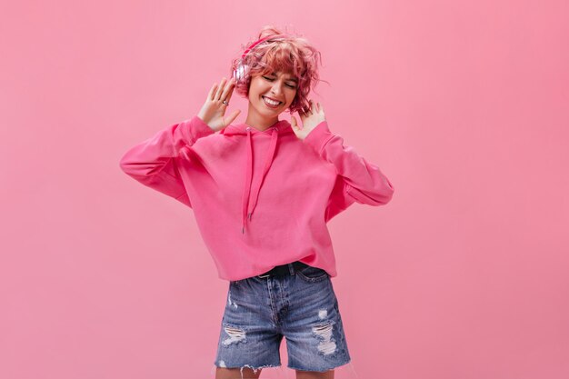 Mulher alegre de cabelo rosa em shorts jeans e capuz enorme ouvindo música em fones de ouvido e dançando na parede isolada