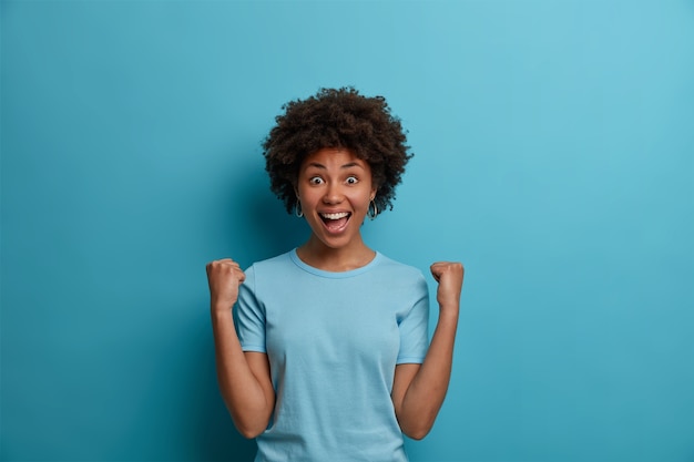 Foto grátis mulher alegre de cabelos cacheados se regozija com a grande chance, bate o punho, comemora excelentes notícias, triunfa sobre algo muito bom, sorri com alegria, encara com os olhos cheios de felicidade, poses internas