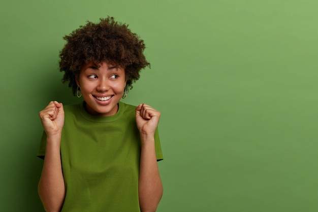 Foto grátis mulher alegre de pele escura com penteado afro, cerra os punhos, aguarda resultados positivos, olha alegremente à parte, vestida com roupas casuais, isolada na parede verde, copie o espaço para seu anúncio