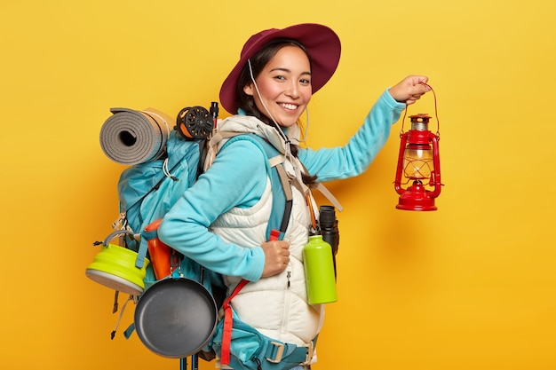 Mulher alegre de trekker segura lâmpada de querosene, usa chapéu e roupa casual, vai descansar na floresta e carrega mochila