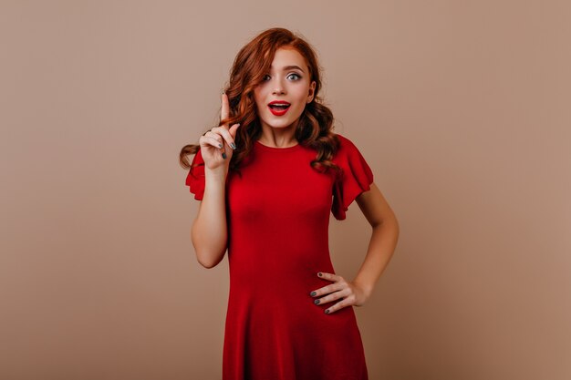 Mulher alegre e bem vestida com cabelo ruivo expressando emoções sinceras. Garota bem humorada em um vestido branco posando.