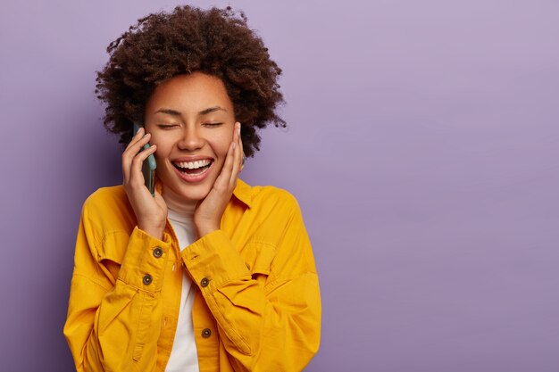 Mulher alegre e despreocupada com penteado encaracolado conversa engraçada com o melhor amigo via smartphone, mantém os olhos fechados e sorri amplamente