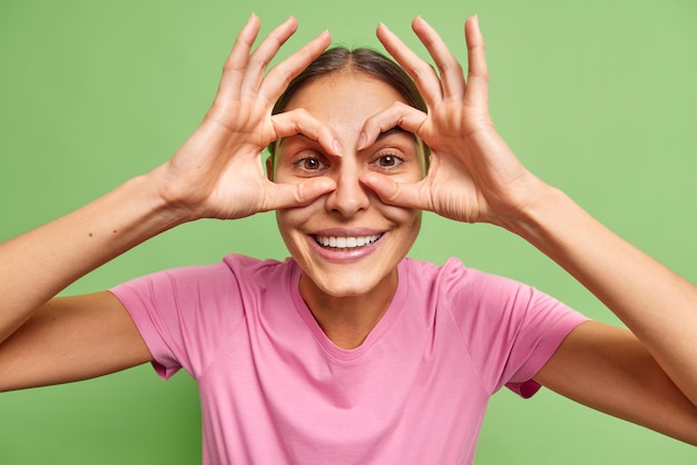 Foto grátis mulher alegre e despreocupada olhando através de círculos faz óculos brincando sorrisos alegremente expressando otimismo veste poses casuais de camiseta rosa contra parede verde vívida