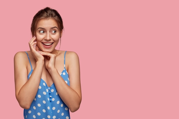 Mulher alegre e emotiva fofa com sorriso cheio de dentes, mantém as mãos perto do rosto, parece com expressão feliz, vestida com vestido de bolinhas, encosta na parede rosa, espaço livre para seu conteúdo promocional