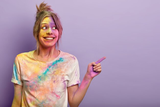 Mulher alegre e encantada brinca com as cores do holi, se diverte no festival, aponta com o dedo indicador, anuncia o espaço da cópia, gosta de cores vivas salpicadas no rosto e na camiseta, faz gestos sobre a parede roxa