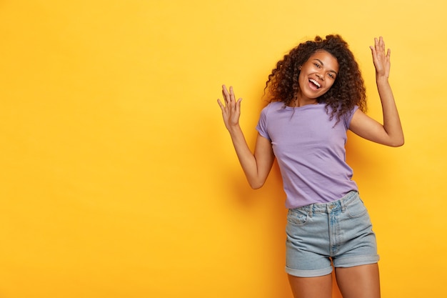 Mulher alegre e otimista de cabelos cacheados levanta os braços, fica radiante, dança ativamente, se diverte na festa, usa camiseta roxa casual e shorts jeans