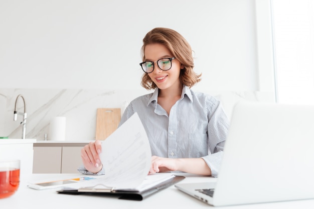 Mulher alegre em copos lendo novo contrato enquanto trabalhava na cozinha