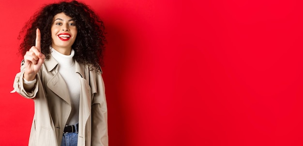 Foto grátis mulher alegre em gabardine mostrando o dedo número um e sorrindo em pé sobre fundo vermelho