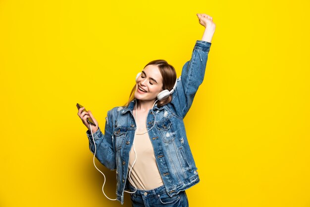 Mulher alegre feliz usando fones de ouvido, ouvindo música de estúdio de smartphone tiro isolado na parede amarela