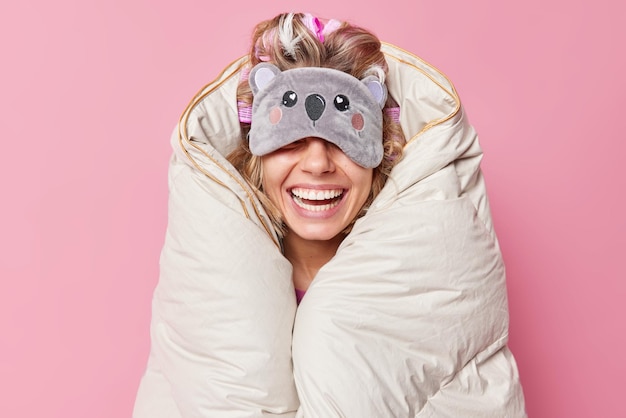 Foto grátis mulher alegre irreconhecível usa máscara de dormir nos olhos sorri amplamente envolto em edredom expressa emoções positivas de bom humor isolado sobre fundo rosa pessoas descansam e conceito de hora de dormir