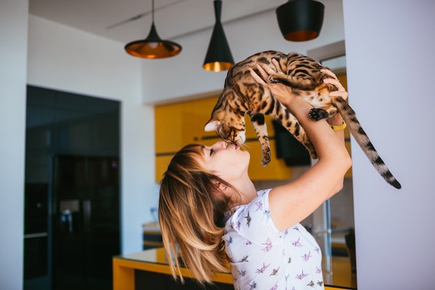 Mulher alegre levanta o gato de Bengala de pé na cozinha