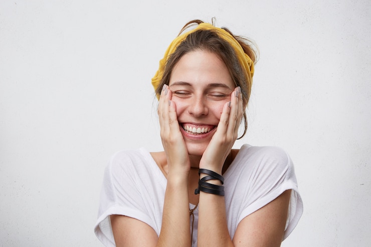Foto grátis mulher alegre positiva segurando as mãos no rosto e sorrindo amplamente fechando os olhos com prazer e bom humor