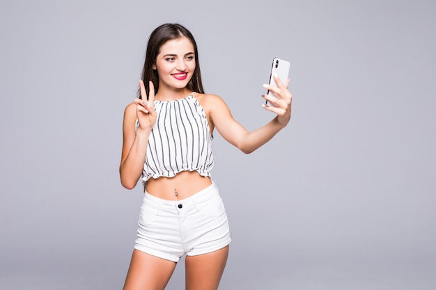 Mulher alegre, sorrindo com dentes brancos e fazendo selfie fotografando a si mesma, mostrando sinal de paz sobre fundo cinza