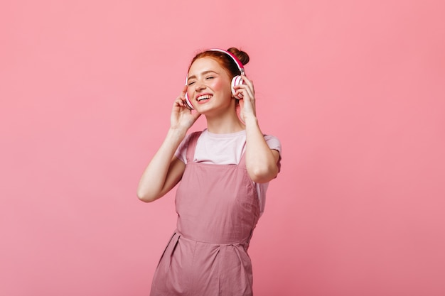 Mulher alegre vestida de macacão leve, dançando e ouvindo música em fones de ouvido no fundo rosa.