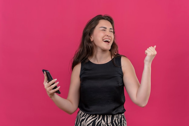 Mulher alegre vestindo camiseta preta e ouvindo música no telefone na parede rosa