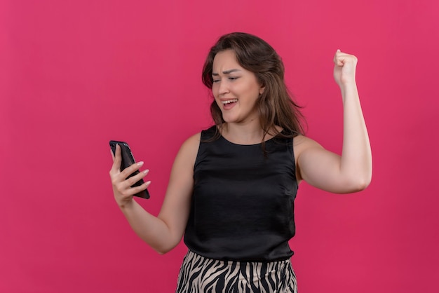 Mulher alegre vestindo camiseta preta ouvindo música no telefone e dançando na parede rosa