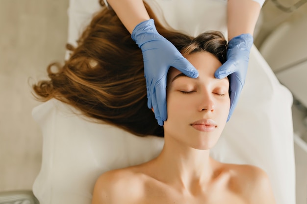 Mulher alegre vista de cima, com longos cabelos castanhos relaxantes da massagem na cabeça de cosmetologista profissional. Tempo para beleza, saúde, rejuvenescimento