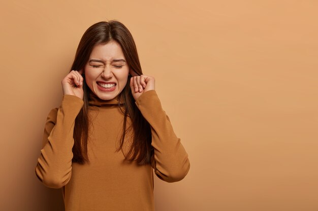 Mulher angustiada e frustrada tapa os ouvidos, cerra os dentes, ouve ruídos incômodos, evita sons desagradáveis, usa roupas marrons, não consegue ouvir mais