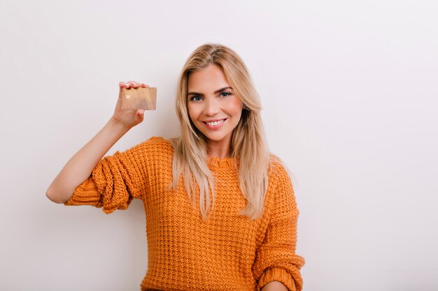 Mulher animada de olhos azuis em um macacão de malha segurando um cartão do banco dourado e rindo