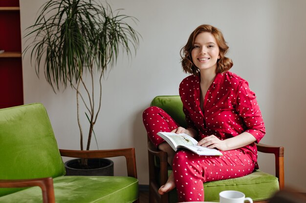 Mulher animada de pijama, sentada na poltrona e olhando para a câmera. Tiro interno de livro de exploração de mulher feliz.