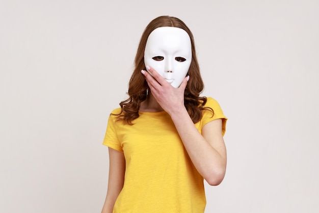 Mulher anônima desconhecida em camiseta amarela estilo casual cobrindo o rosto com máscara branca, escondendo personalidade, conspiração e privacidade. tiro de estúdio interior isolado em fundo cinza.