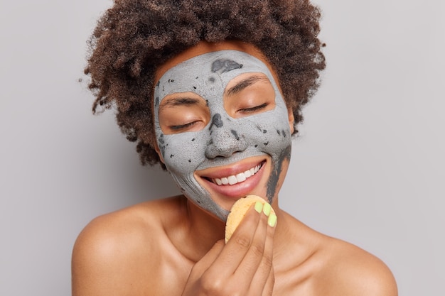 Mulher aplica máscara de argila com esponja cosmética sorri gentilmente gosta de procedimentos de cuidados com a pele fecha os olhos de poses de prazer nua dentro de casa. conceito de beleza.