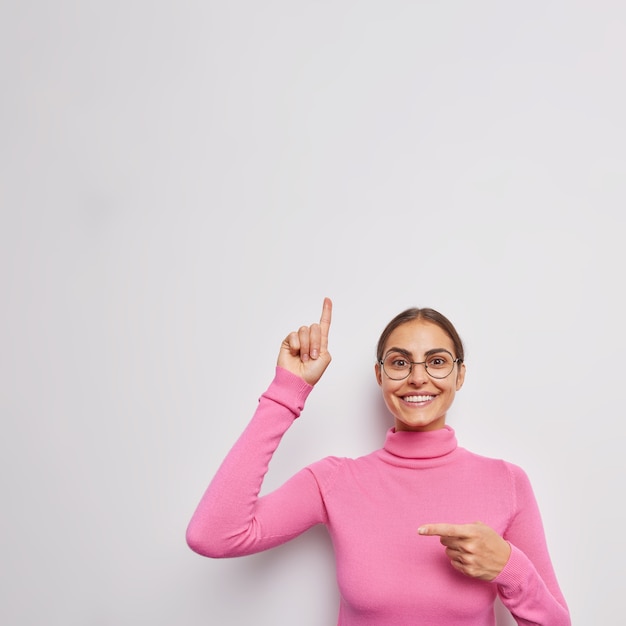 Foto grátis mulher aponta para cima e sorri de lado mostra gentilmente a direção ou o caminho para algum lugar vestido com um poloneck casual em branco