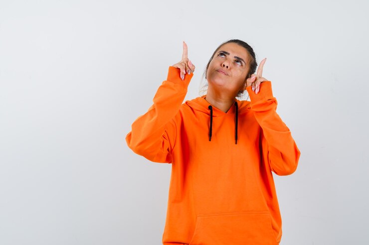 Foto grátis mulher apontando para cima com um capuz laranja e parecendo esperançosa