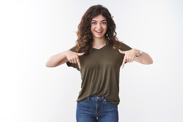 Foto grátis mulher armênia confiante, jovem e atrevida, com cabelo encaracolado, vestindo camiseta sorrindo emocionada, você vai gostar expressão apontando para baixo os dedos indicadores mostram um produto legal promocional interessante