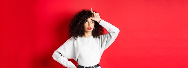 Foto grátis mulher arrogante com cabelo encaracolado mostrando sinal de perdedor na testa e olhando de lado zombando de alguém stan